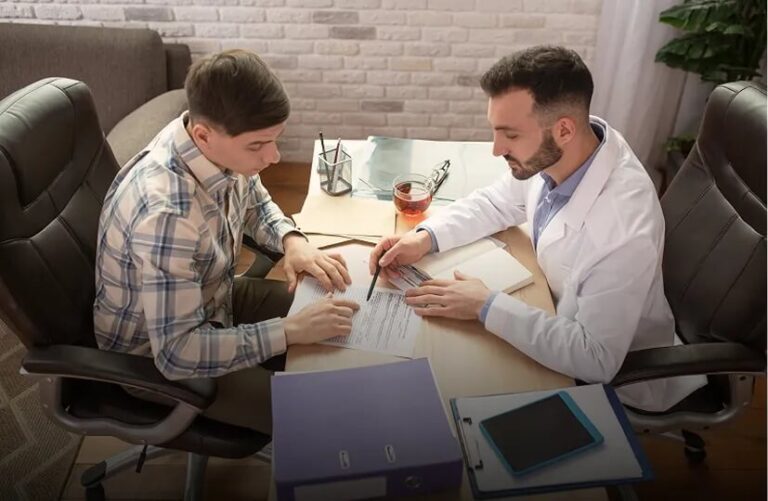 Na zdjęciu lekarz w białym fartuchu omawia z pacjentem dokumentację medyczną podczas konsultacji. Mężczyźni siedzą przy biurku, analizując wyniki i dokumenty.